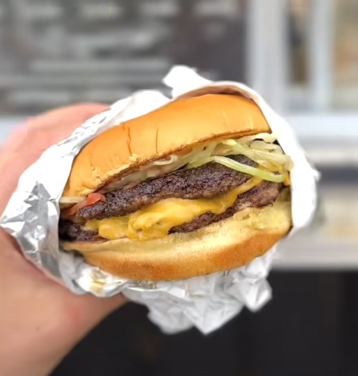 Hand-held double smash burger wrapped in foil, showing melted cheese, fresh lettuce, tomato, and secret sauce